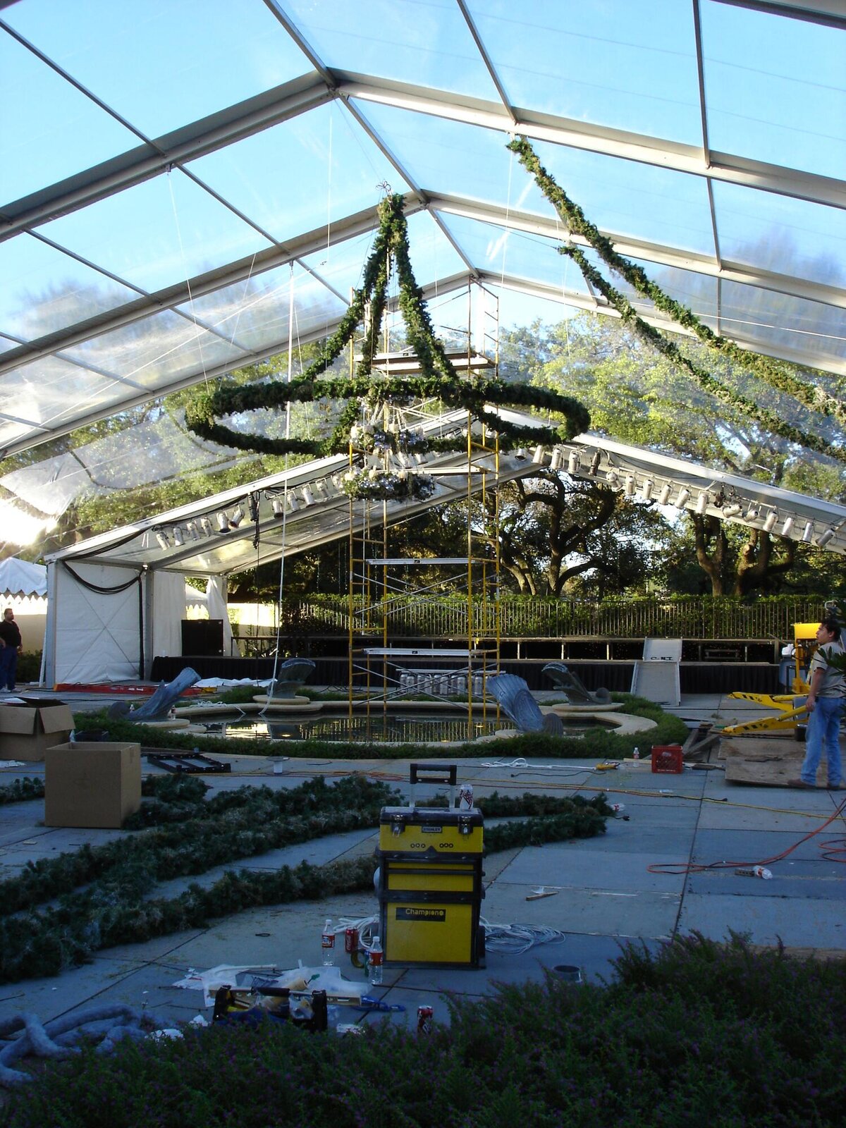 Clear-top tent during build — chandelier rig and greenery garlands rising over the estate pool.